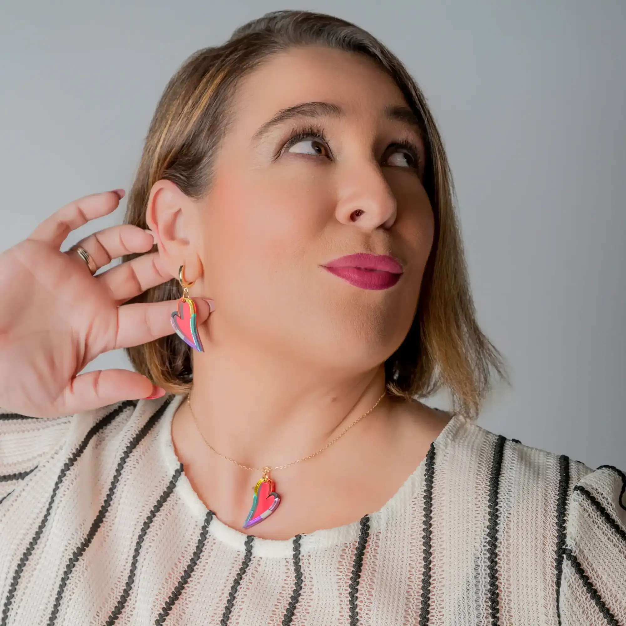 Modelo con el colgante Latido en forma de corazón alargado, en color arcoíris, con cadena chapada en oro de 14k. Plano beauty donde también se aprecian los charms combinados con la joya.